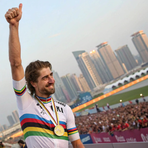 Peter Sagan, celebrating a victory. Joyful face, wearing a white and color striped jersey with a gold medal. Raised thumb. Behind him, a crowded stadium and tall buildings of Doha, Qatar