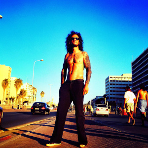 George Lincoln D'Costa, known by his stage name Artcell, stands out with a fierce look. He sports a muscular build, curly dark hair, and distinctive tattoos. Wearing glasses. Midday in a bustling city street.