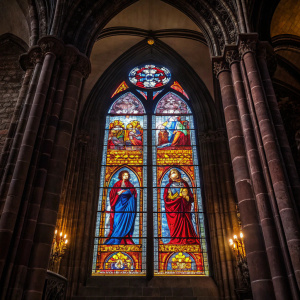 Stained glass window from 14th-century Strasbourg Cathedral. Vibrant colors: red, blue, and yellow depict religious figures. Dark gothic cathedral interior as backdrop