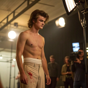 Joe Keery, shirtless, with a bandage around his waist. Blood visible. Behind-the-scenes of Stranger Things. Studio lights, blurred crew in background