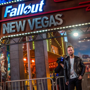 Aaron Paul at the E3 gaming convention. Promotional backdrop featuring 'Fallout: New Vegas'. He wears a black leather jacket over a white shirt.