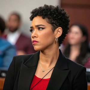 Alexandra Shipp, African-American actress, with short curly hair, frowning intensely. She is wearing a black blazer and red top. At a press conference with blurred attendees in the background.