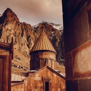 Monumental Geghard Monastery, Kotayk Province, Armenia. Stone architecture, dome-shaped buildings, reddish-brown hues. Surrounded by rugged mountains with snow-capped peaks and ancient stone walls.