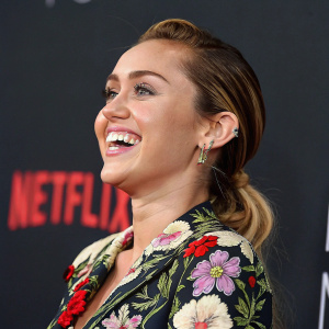 Close-up of Miley Cyrus at Netflix event. Smiling broadly. Wearing a floral embroidered top. Solid black background