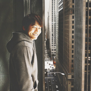 Son Ho-jun, Asian actor, stands in front of a large cityscape. Wearing a casual grey hoodie, smiling at the camera.