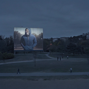 Robinson Cano at a bustling city park, wearing a casual hoodie and jeans. Grinning broadly, standing in front of a towering billboard featuring a Nike advertisement.
