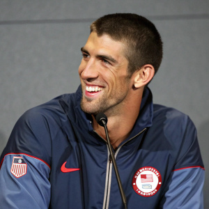 Michael Phelps, smiling confidently, wearing an olympic jacket with NCI patch. Indoor press conference. Simple gray backdrop.