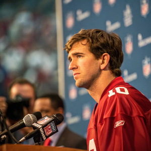 Eli Manning, serious face, wearing a red jersey. At a press conference for a football game. The background features a grid of microphones and a blurred audience.