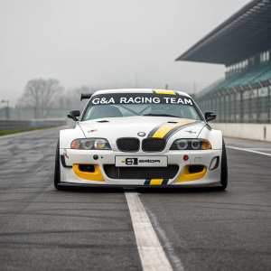 A sleek BMW Z3 M Coupe GT race car, painted white with black accents and a vibrant yellow stripe. The car is emblazoned with logos from the G&A Racing Team. Shown on a desolate gray track.