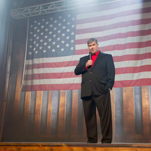 Sean Hannity, conservative talk show host, standing on a stage. The backdrop is an American flag. He's dressed in a black suit with a red shirt, holding a microphone.