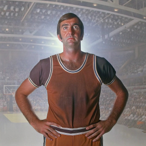 Gail Goodrich, former basketball player, posing in his classic Lakers jersey. Indoor arena setting with a crowd blurred in the background.