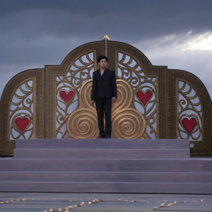 Shah Rukh Khan in a black suit, standing in front of a grand stage with intricate golden designs and red heart-shaped motifs. He appears serious and powerful, the golden backdrop enhancing his presence.