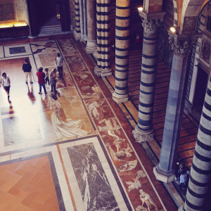 A stunning panoramic view of the Siena Cathedral's marble mosaic floor. Rich hues of brown, tan, and gray intertwine in detailed patterns. Historical scenes depicted amidst towering columns and arches, adorned with golden accents. Visitors wandering through the sacred space.