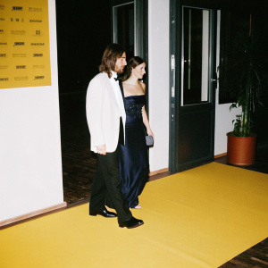 Sharlto Copley and Tanit Phoenix attending the Berlin Film Festival. Formal attire and relaxed hairstyle. Dark blue gown and neat coiffure. Red carpeted area, sponsor logos backdrop.
