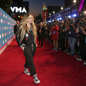 Avril Lavigne on the red carpet at the MTV Video Music Awards. She is dressed in a casual yet chic outfit, with her signature long, wavy hair. The scene is bustling with fans and other celebrities, the backdrop filled with vibrant VMA logos.