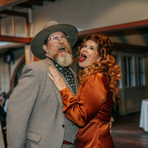 Elliott Gould and Jennifer Bogart, a couple posing for a candid shot at a vintage indoor gala. Gould, with his signature large brimmed hat and scruffy beard, is energetically positioned next to Bogart, whose hair is in luxurious curls. The dimly lit venue with blurred details in the background enhances the vintage feel.
