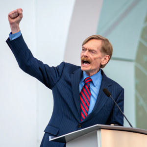 Robert Zoellick, in a vibrant keynote speech. Dynamic pose with podium, dark blue pinstripe suit, blue shirt, and red-striped tie. Minimalist, muted background