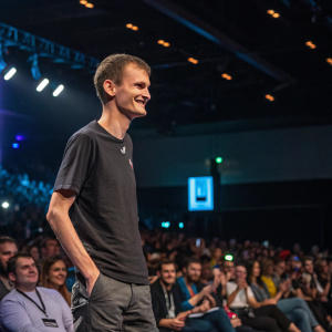 Vitalik Buterin smiling on stage. Tech conference with rows of attendees in the background.