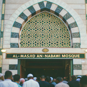 Archway entrance of Al-Masjid an-Nabawi Mosque. Detailed geometric patterns and Arabic script carved into light-colored stone. Crowd of people in traditional Muslim attire.