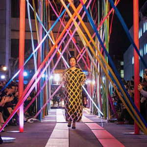 Jung Kook at the fashion week runway show. Designer wear, geometric pattern dress. Runway surrounded by cheering crowd.