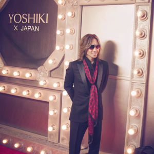 Yoshiki, musician from X Japan, at a Hollywood premiere. Black tuxedo, red and black patterned scarf. Confident pose. Event logo backdrop, bright lights.