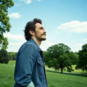 James Franco, mustache and beard, in a casual stance. Blue sky background, green park. Wearing a denim jacket.