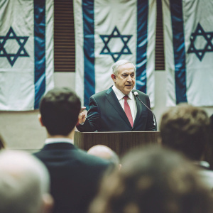 Benjamin Netanyahu, in a formal navy suit and a red tie, addressing a room full of people at the Knesset. Blue and white Israeli flags prominently displayed behind him.