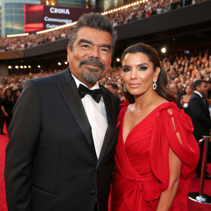 George Lopez and Eva Longoria at a Hollywood Awards Show. George Lopez, wearing a black tuxedo, Eva Longoria in a flowing red dress. Large crowd in the background