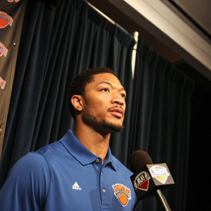 Derrick Rose, in a blue polo shirt with New York Knicks logo, center stage at a press conference. Dark backdrop with draped curtains. Microphone with Knicks logo tag in foreground. Close-up photo.
