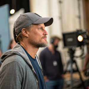 Woody Harrelson, in a casual outfit with gray cap and beard. Staring intently at something, candid shot in a blurred studio environment.