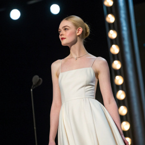 Close-up shot of Elle Fanning at award show. Wearing minimalist white dress. Dark stage backdrop, spotlight on her