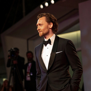 Tom Hiddleston, dressed in a tuxedo, at the Venice Film Festival. Poses confidently on a dimly lit stage.