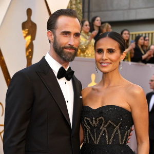 Joseph Fiennes and Maria Dolores Dieguez, glamorous attire, at a prestigious film gala. Black strapless gown with intricate beadwork, classic tuxedo, bow tie, against backdrop of golden statues and applauding audience.