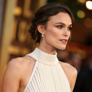 Close-up photo of Keira Knightley. Wearing a high-neck, sleeveless, ribbed white gown. Background blurred with warm, golden hues.
