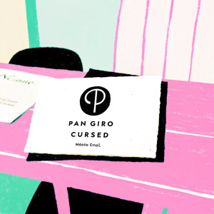 Business card for Pan Giro's 'Cursed' brand. Modern abstract logo. White card with black text. Website and email details. Laid on a wooden desk in a minimalist office space.
