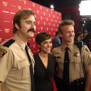 Reno 911! cast in action. Man with mustache, woman with short haircut, and another man with similar hairstyle in tan police uniforms. Behind the scenes at Spike TV awards. Red backdrop with Spike TV logo.