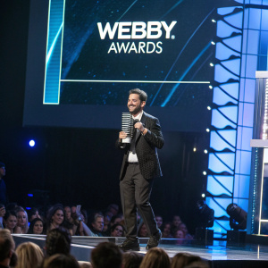 Man accepting award at Webby Awards ceremony. Large screen displaying award category behind him.