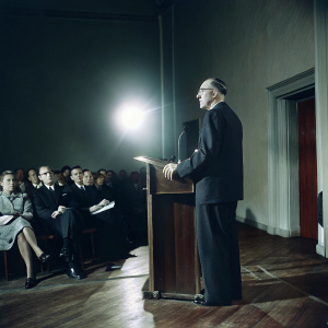 Sir Menzies Campbell delivering a speech. Dimly lit auditorium with audience seated. Bright stage lights