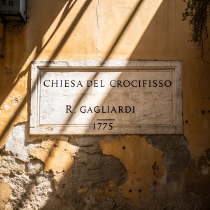 An ancient wall adorned with a weathered Italian inscription. The text reads 'Chiesa del Crocifisso' and 'R. Gagliardi', dated 1715. The wall is exposed to bright sunlight.