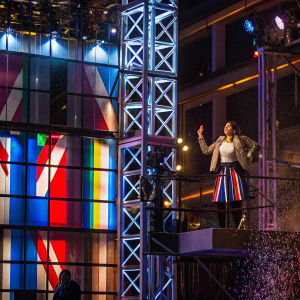Dua Lipa, the pop sensation, on stage performing. Wearing a white blouse, leopard print jacket, and Union Jack skirt. Stage lights with colorful patterns. Confetti-like particles in the air.