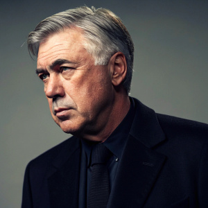 Close-up of Carlo Ancelotti, Italian football manager. Serious face. Dark suit. Soft lighting highlights his features and attire. Muted background.