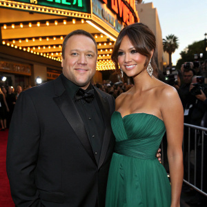 Kevin James and Steffiana de la Cruz attending the premiere of 'Zookeeper'. They are both dressed formally, Kevin in a black tuxedo, Steffiana in a strapless emerald gown. They are standing in front of a brightly lit marquee, surrounded by flashing cameras and curious onlookers.