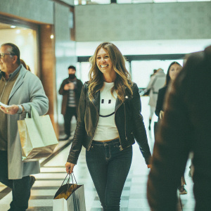 Elizabeth Hurley at a bustling indoor mall, wearing a black leather jacket, dark jeans, and a white t-shirt with a smiley face. She's pushing a designer shopping bag and pausing for a photo.