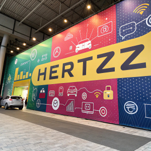 Hertz banner in vibrant English script at a tech conference. Features car rental offer, digital icons, and eye-catching design.