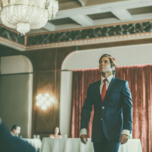 Cory Feldman, actor, in a sleek navy suit with red tie. At a gala event. Luxurious ballroom with chandeliers and velvet red curtains.