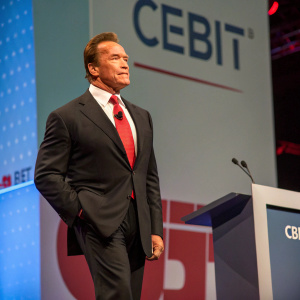 Arnold Schwarzenegger at a tech conference. Dark suit, red tie. CEBIT logo backdrop