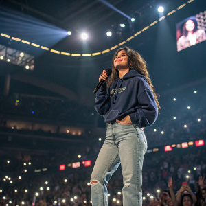 Camila Cabello at a concert. She wears a casual outfit, jeans and a hoodie. The venue is brightly lit with fans in the background.