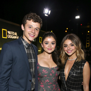 Nolan Gould, Ariel Winter, and Sarah Hyland, smiling, posing for a photograph. Dark stage with bright lights and National Geographic banners in the background.