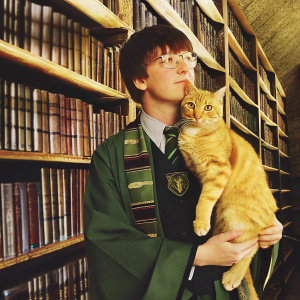 Draco Malfoy, a young wizard, with a ginger cat. Green robes, Slytherin crest, standing in a library with tall, shadowy shelves.