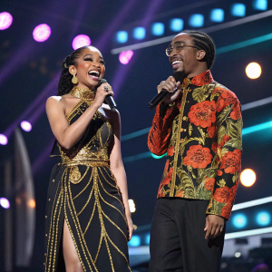 Saweetie and Quavo on stage at Grammy Awards. Saweetie in a black and gold, traditional Chinese-inspired gown. Quavo in a patterned shirt. Cheerful pose, colorful stage lights